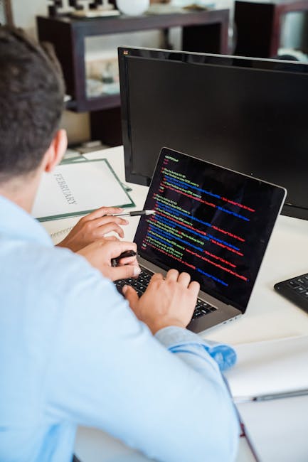 A person with short dark hair, wearing a light blue shirt, is seated at a desk outdoors, working on a laptop displaying colorful lines of code. The workspace includes a black monitor, a notebook, and 
