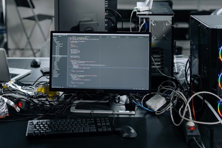 A person with short dark hair, wearing a light blue shirt, is seated at a desk outdoors, working on a laptop displaying colorful lines of code. The workspace includes a black monitor, a notebook, and 