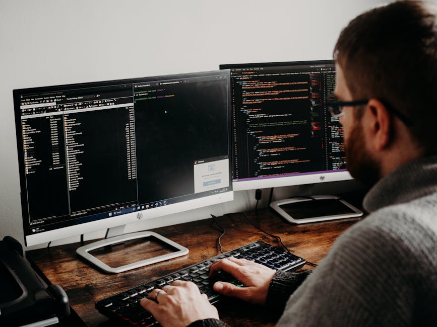 A person with glasses and a beard is sitting at a dark wooden desk, working on a computer with dual monitors. The screens display lines of code and a file directory, indicating programming activity. T