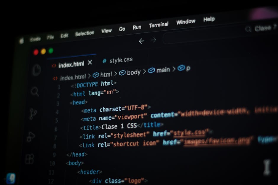 A person with glasses and a beard is sitting at a dark wooden desk, working on a computer with dual monitors. The screens display lines of code and a file directory, indicating programming activity. T