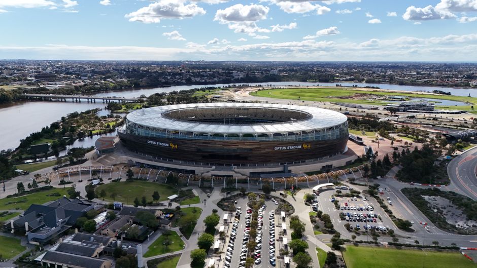 An aerial view of Optus Stadium situated along the Swan River, showcasing its modern, oval-shaped structure with a partially open roof. Surrounding the stadium are meticulously maintained landscaped a